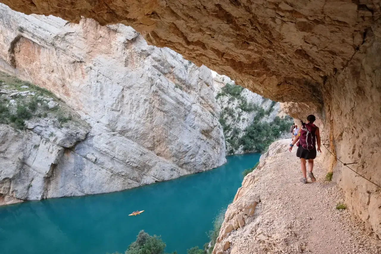 Senderismo por la sierra del Montsec, con un cañón de roca y un río turquesa. Un kayak navega en la distancia.
