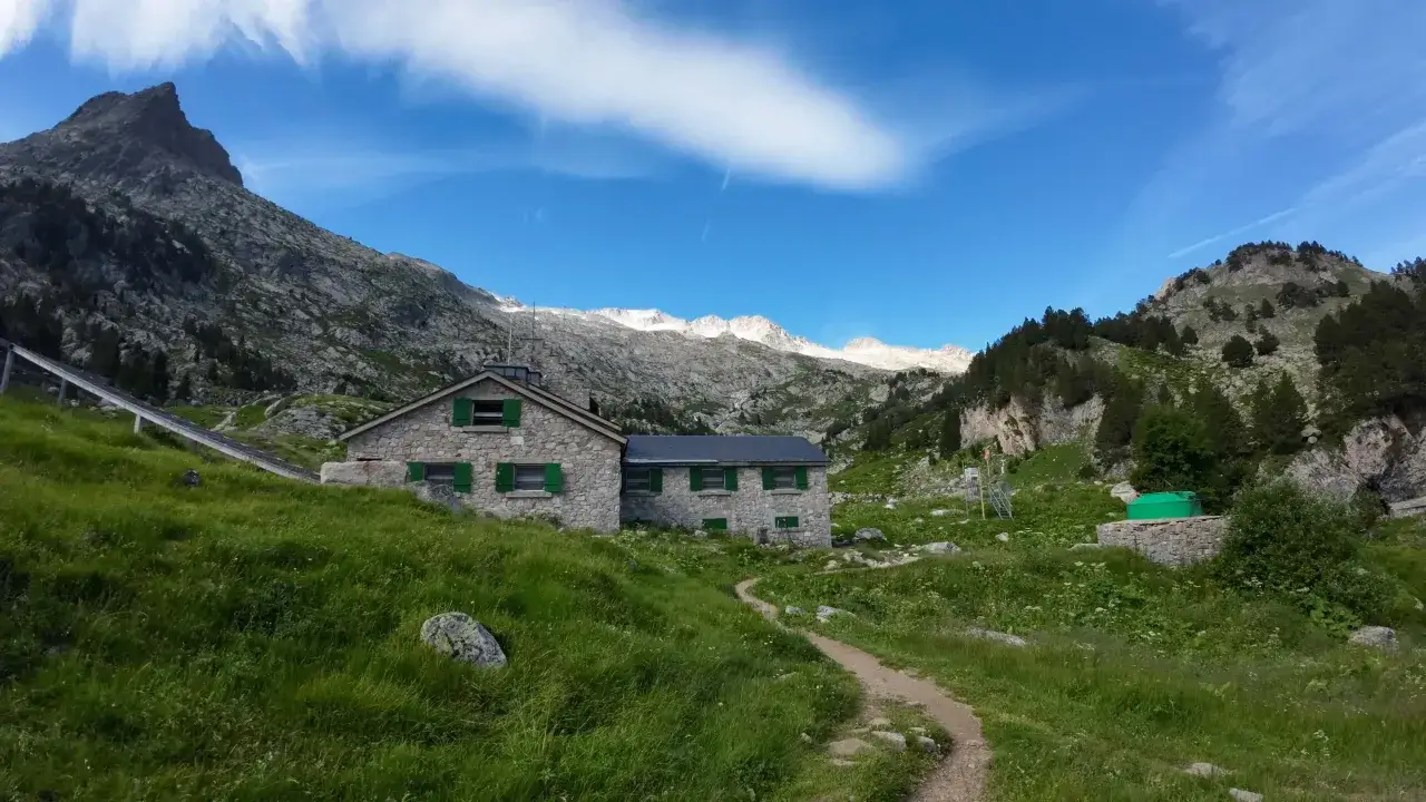 Refugio de montaña de piedra con ventanas verdes, rodeado de prados y picos rocosos. Un sendero serpentea hacia la casa, que parece la