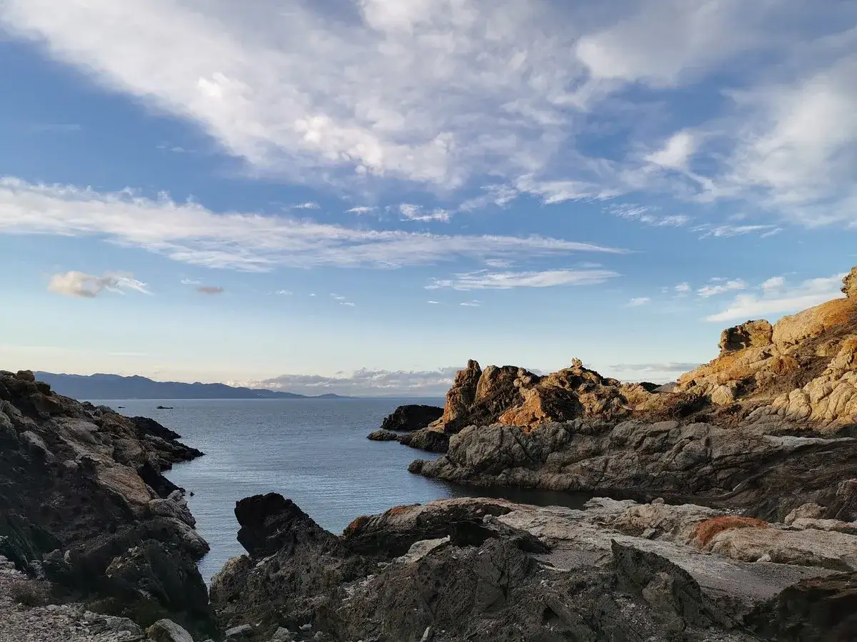 Paisaje rocoso y marino en el paratge de Tudela. El mar azul se abre entre formaciones rocosas doradas bajo un cielo nublado.