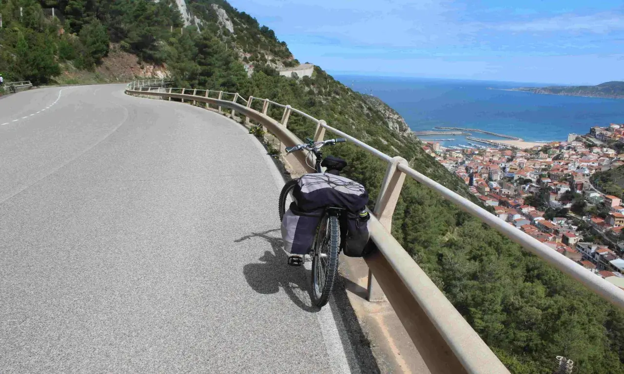 Bicicleta cargada lista para un tour en bicicleta por la costa de Cerdeña, con vistas a un pueblo costero y el mar azul.