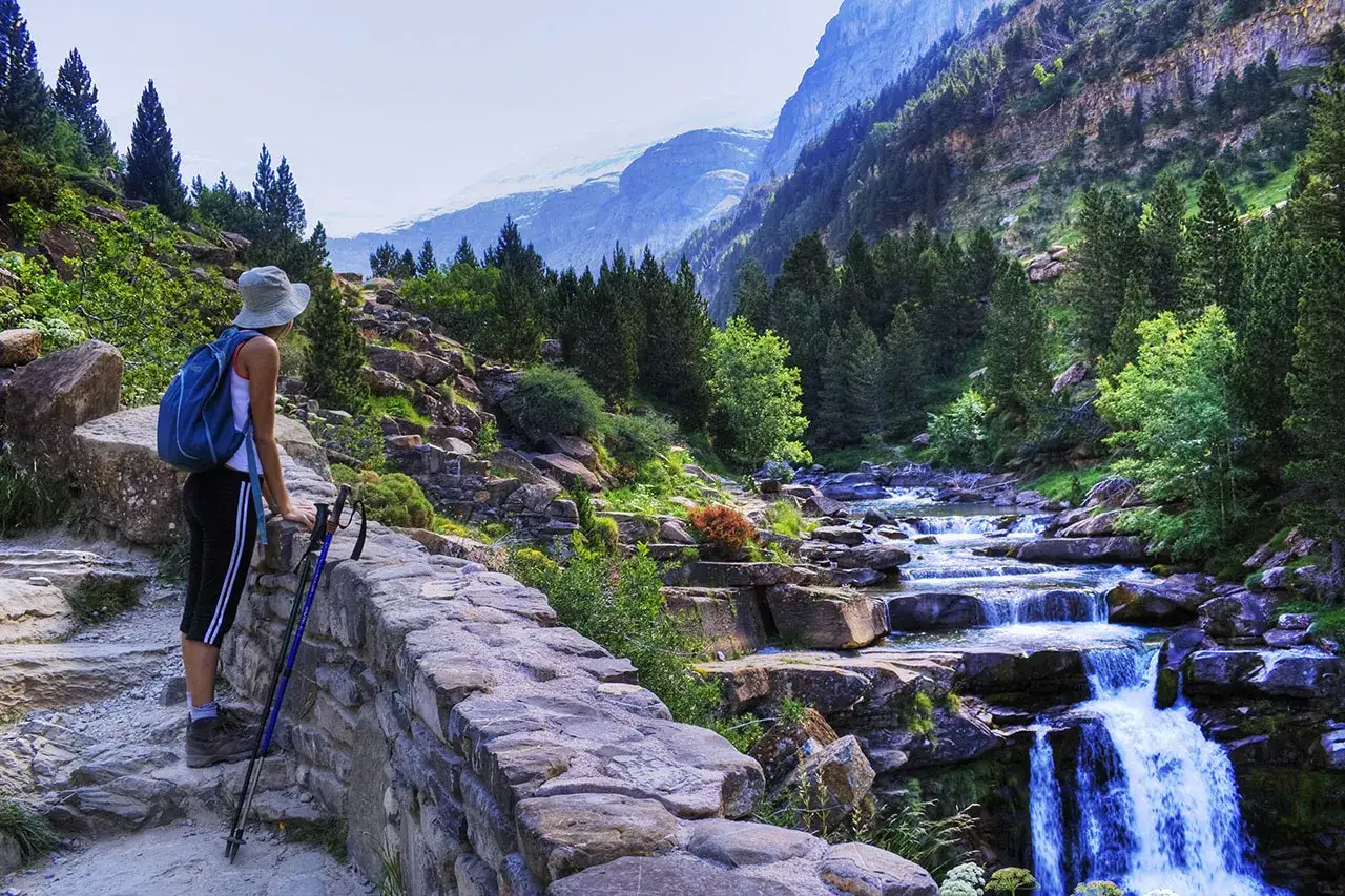 Senderista contempla un río en cascada en los Pirineos de Huesca, rodeado de bosques y montañas.