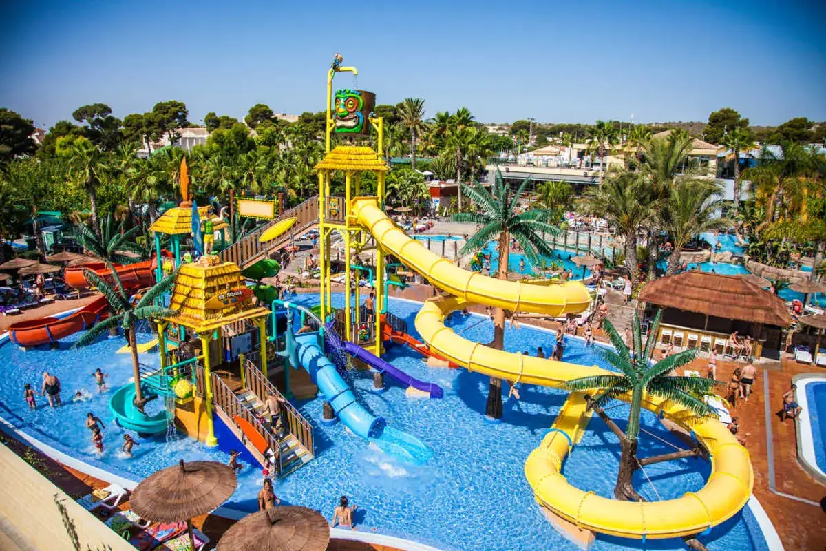 Un parque acuático con toboganes amarillos y azules, palmeras y gente disfrutando del sol. ¡Un lugar ideal para camping con toboganes en España!