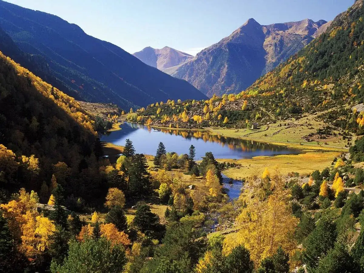 Lago sereno en el Pirineo Catalán, rodeado de montañas y árboles de otoño con hojas doradas.
