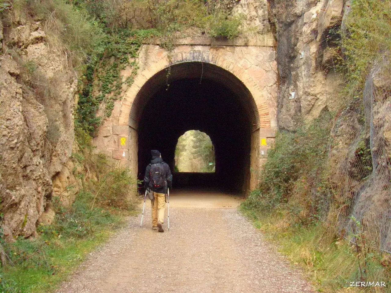 Senderista se dirige al túnel del carrilet Cal Rosal, rodeado de vegetación y rocas.