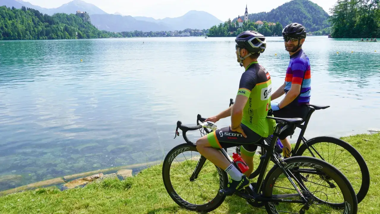 Ciclistas disfrutan de un descanso en sus viajes en bicicleta por Europa, junto a un lago con montañas y un castillo al fondo.