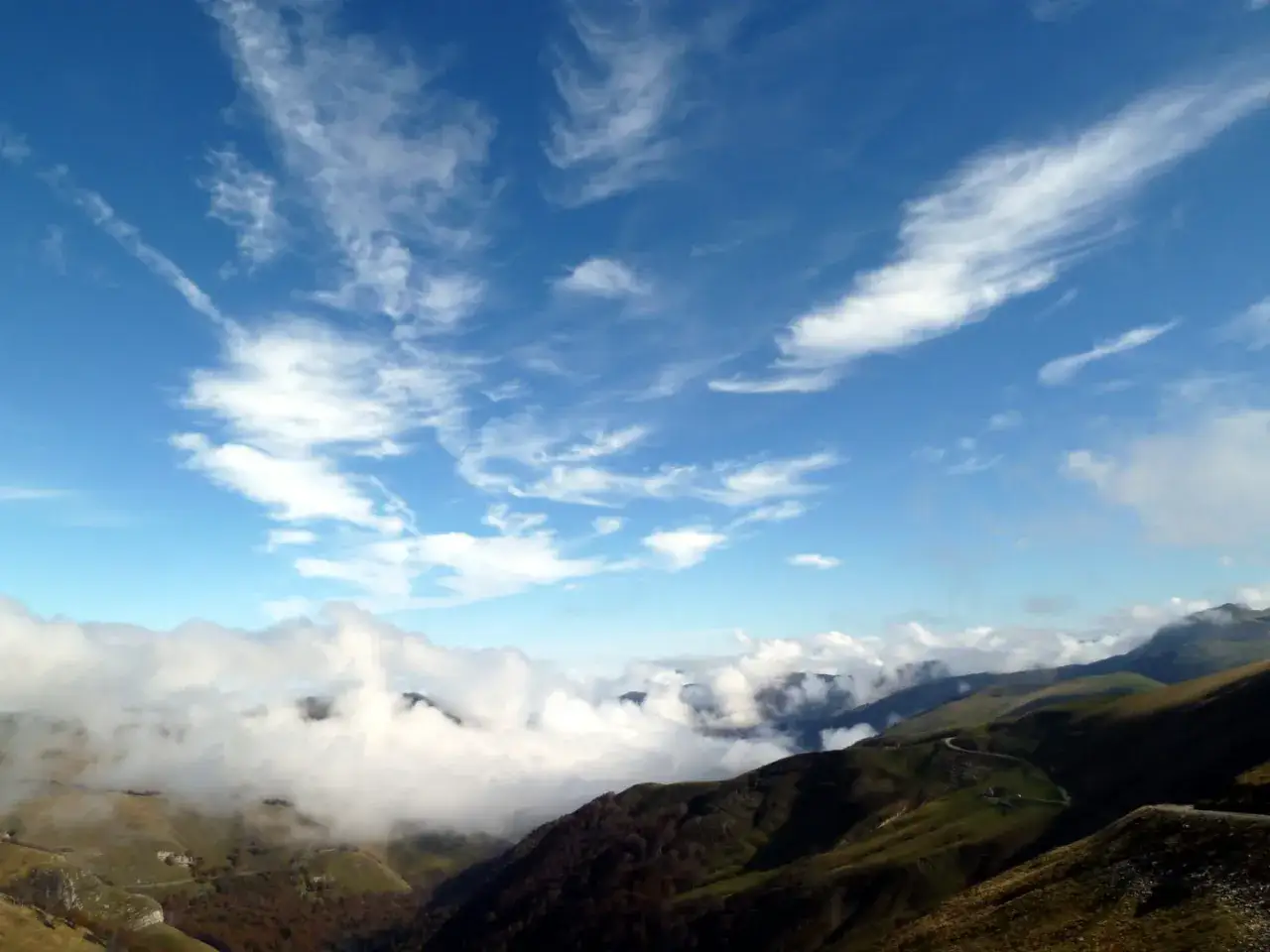 Paisaje montañoso con nubes bajas y cielo azul. Una carretera serpentea por las laderas verdes, evocando la ruta del puerto de Larrau.