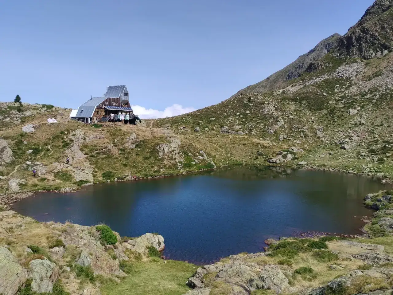 El refugio de Vallferrera se alza junto a un lago alpino, rodeado de montañas rocosas y vegetación de montaña.