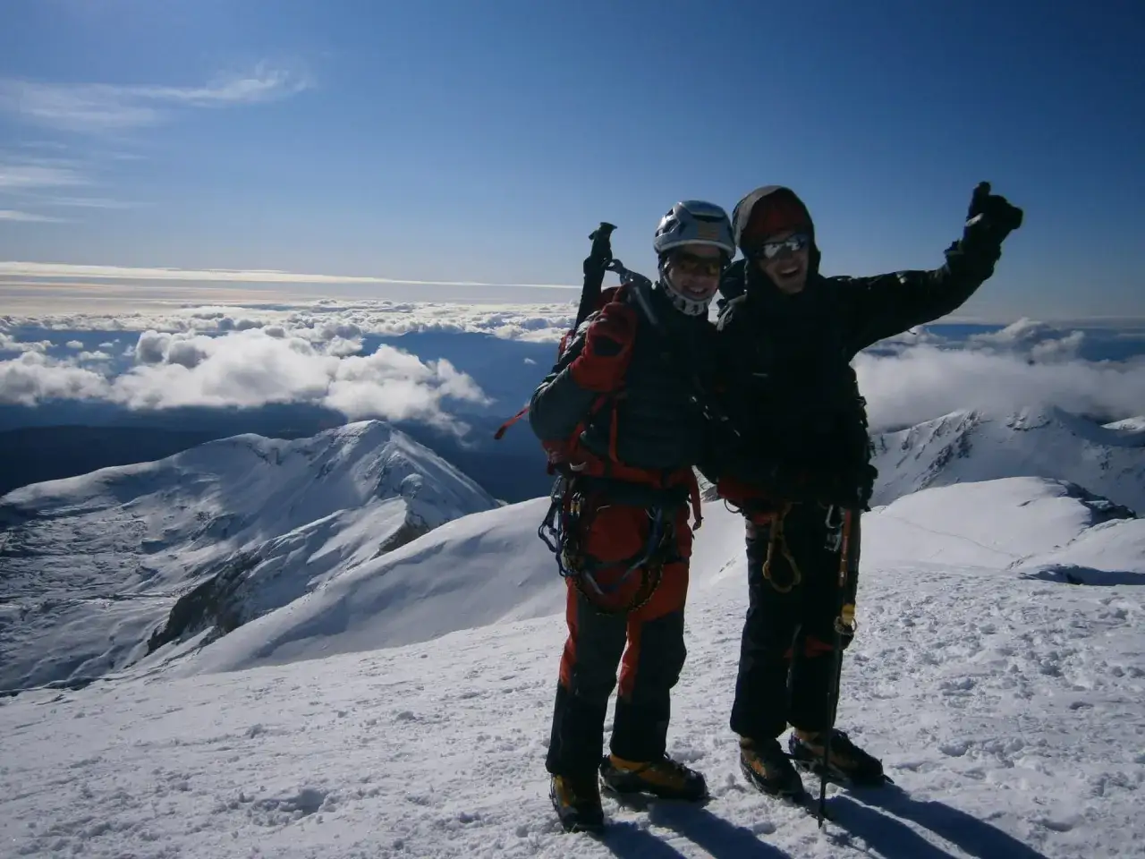 Dos alpinistas celebran en la cima nevada del pico Taillón, con un mar de nubes bajo un cielo azul.