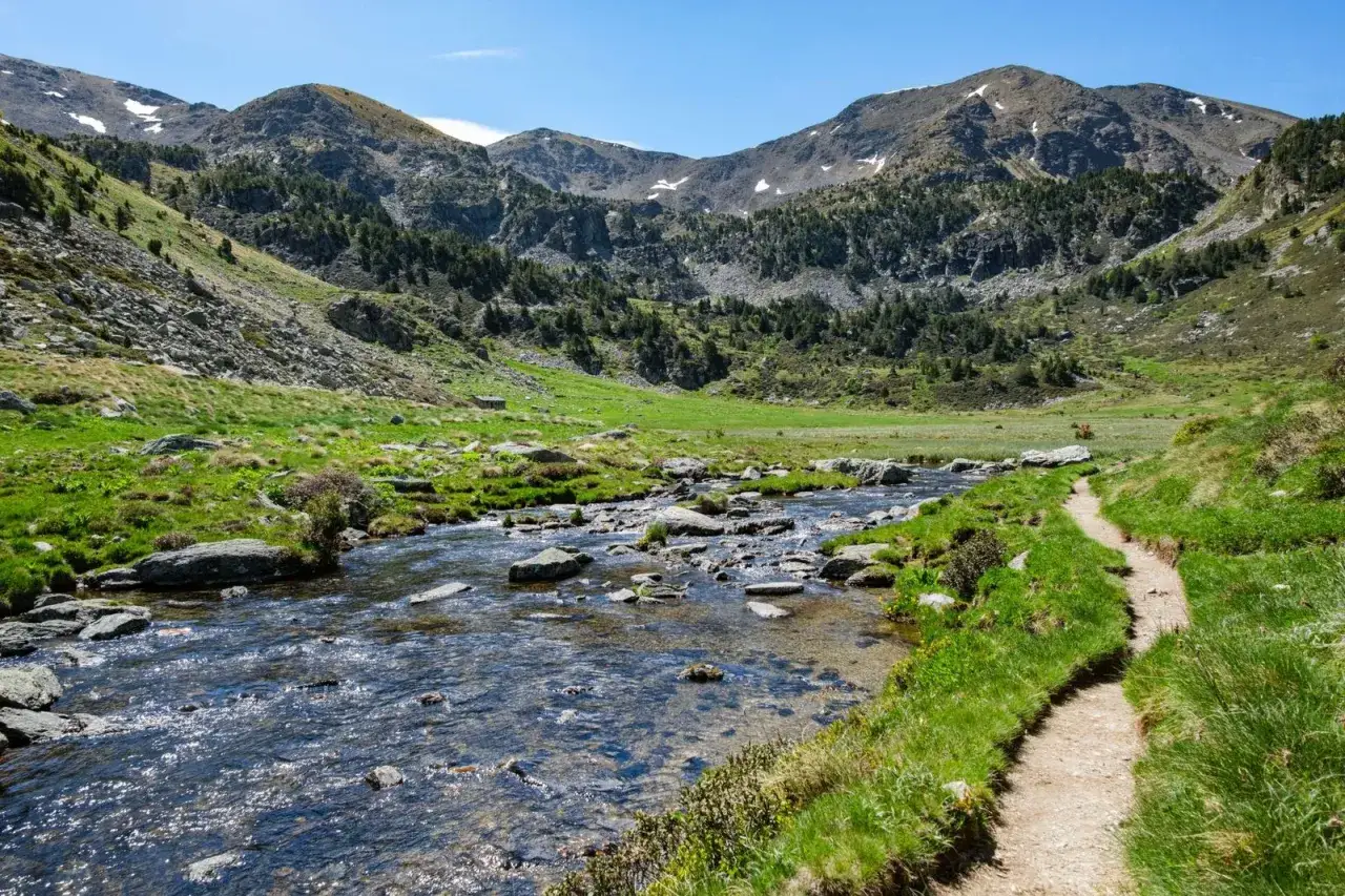 Sendero junto a un río pedregoso en el Vall d'Incles, rodeado de prados verdes y montañas boscosas bajo un cielo azul.