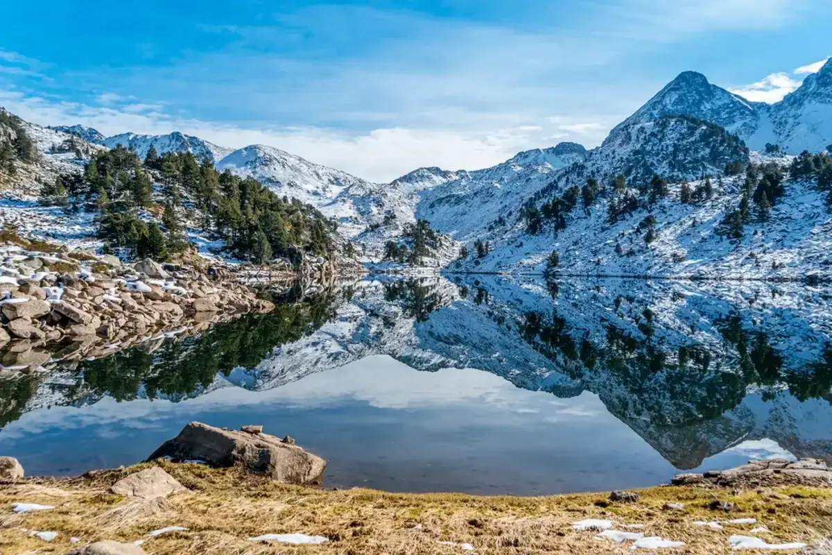 Espectacular reflejo de montañas nevadas y pinos en un lago sereno en el Pla de Beret. El cielo azul contrasta con la nieve y la roca.