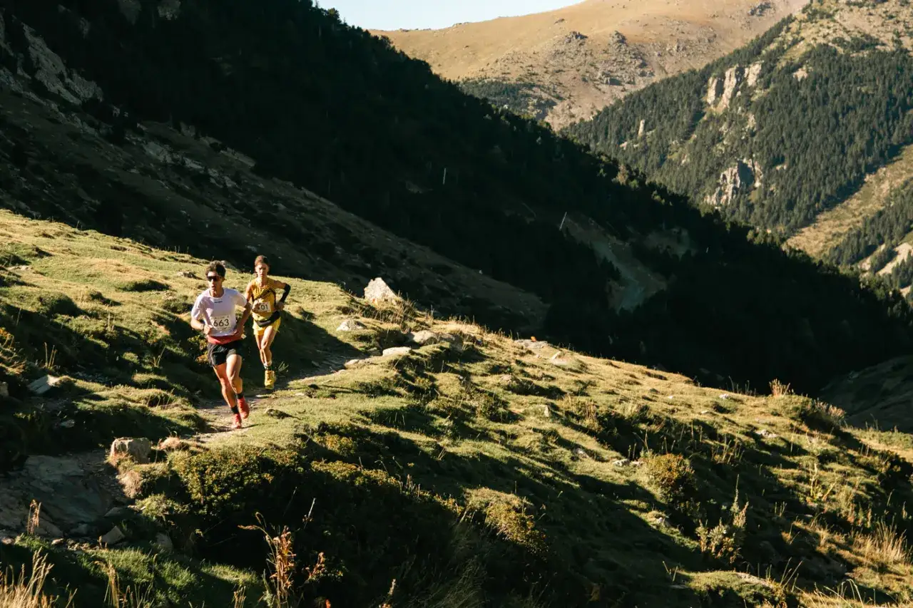Dos corredores compiten en l'olla de Núria, un paisaje montañoso con bosques y laderas verdes bajo un cielo azul.