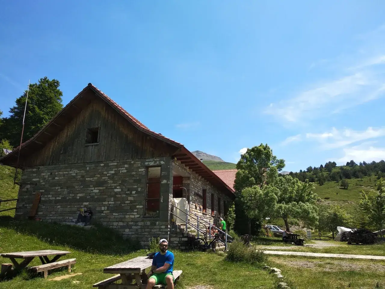 Un hombre descansa en un banco frente al refugio de Linza, un edificio de piedra y madera rodeado de verde bajo un cielo azul.