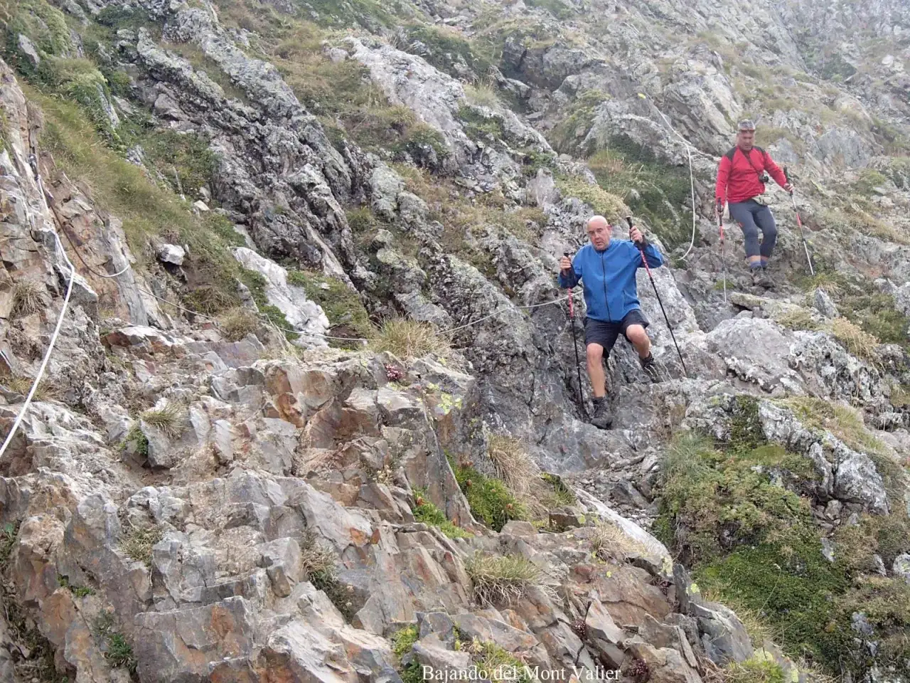 Dos excursionistas descienden por una ladera rocosa y empinada del Mont Valier, usando bastones para mantener el equilibrio.