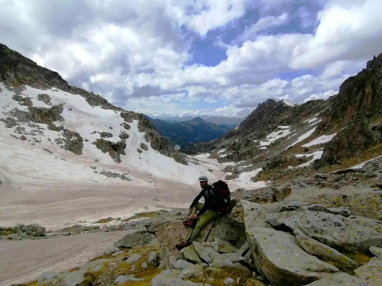 Un excursionista descansa en el valle de Aneu, rodeado de picos rocosos y parches de nieve bajo un cielo nublado.