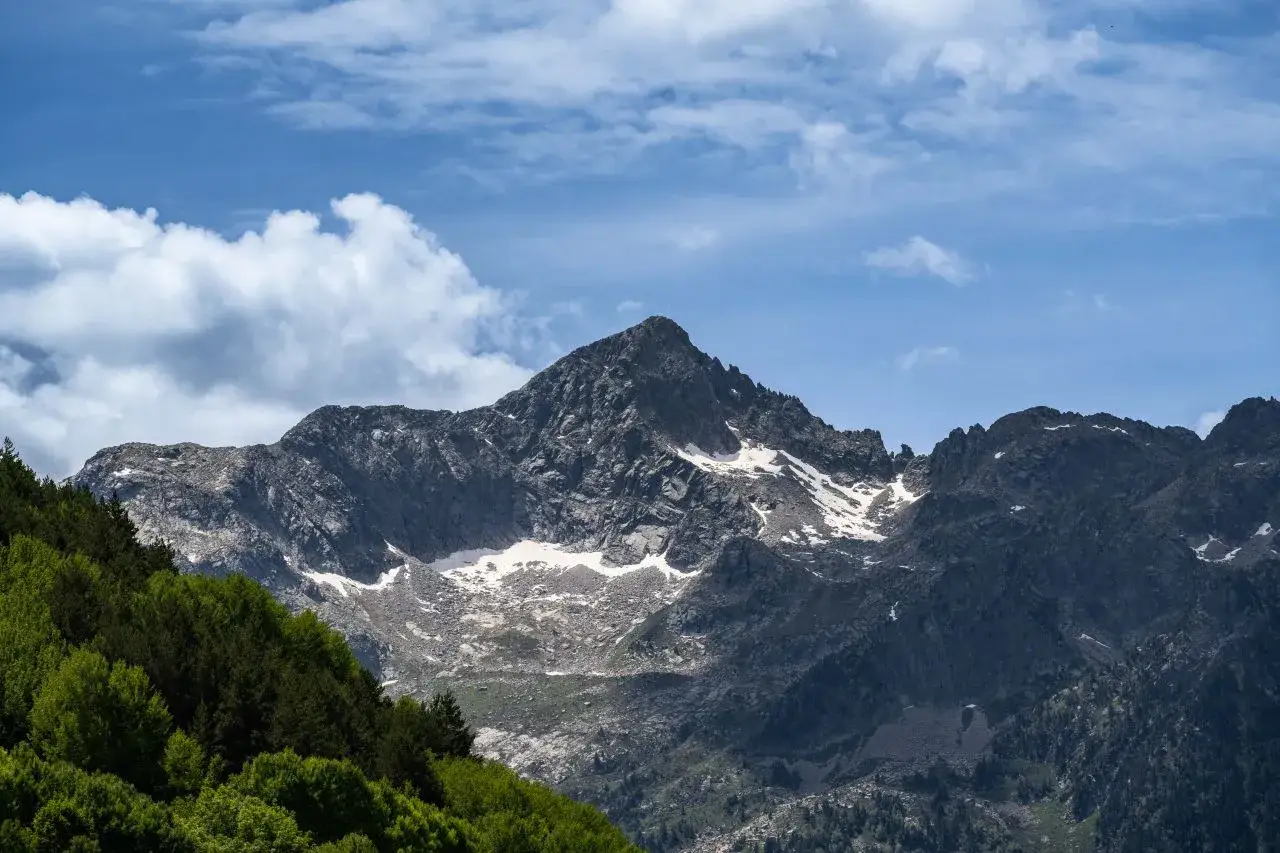 Majestuoso montarto pico rocoso con nieve, rodeado de verdes bosques y cielo azul con nubes.