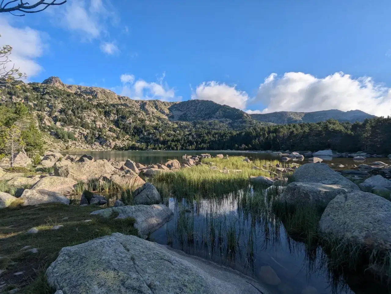 Paisaje sereno junto al refugi de Malniu, con un lago tranquilo reflejando montañas y cielo azul. Rocas y vegetación bordean la orilla.