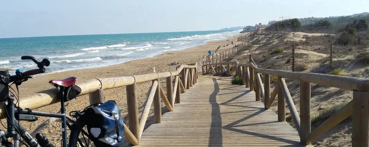 Bicicleta lista para recorrer la ruta EuroVelo 8, con el mar Mediterráneo y una pasarela de madera como escenario.