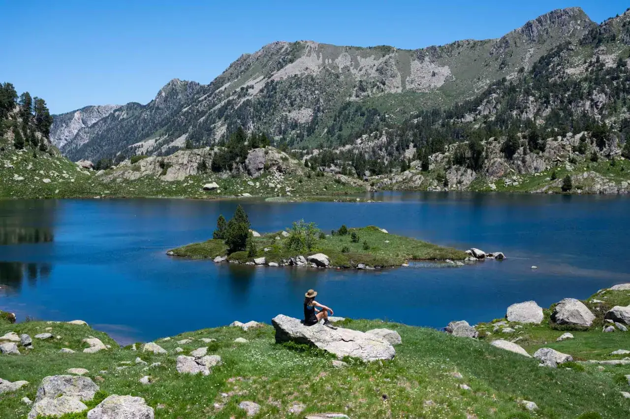 Persona sentada en roca contempla un lago azul rodeado de montañas, ideal para rutas en el Valle de Arán.