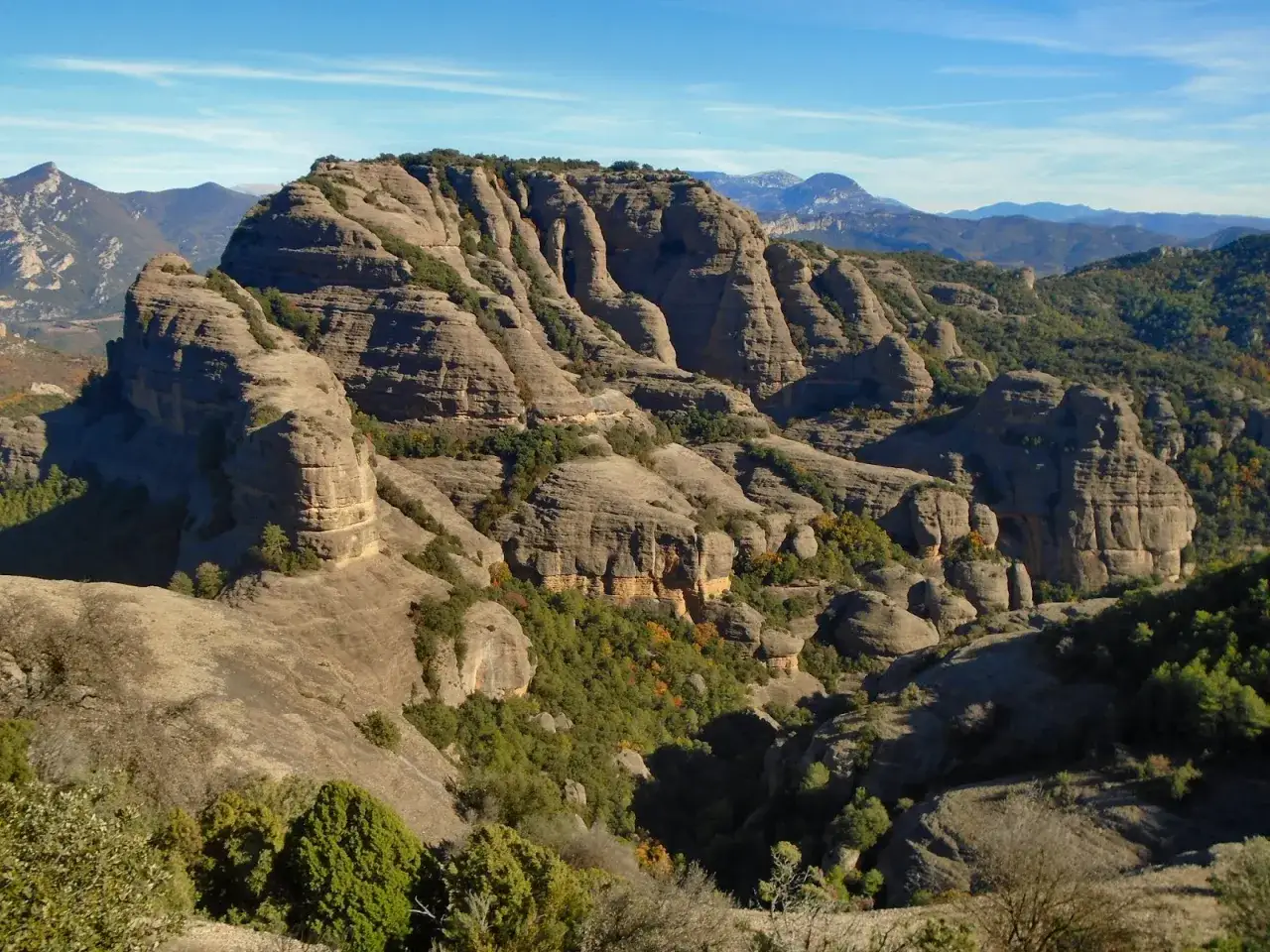 Majestuosa formación rocosa, la Roca del Corb, se alza entre barrancos y vegetación, bajo un cielo azul intenso.