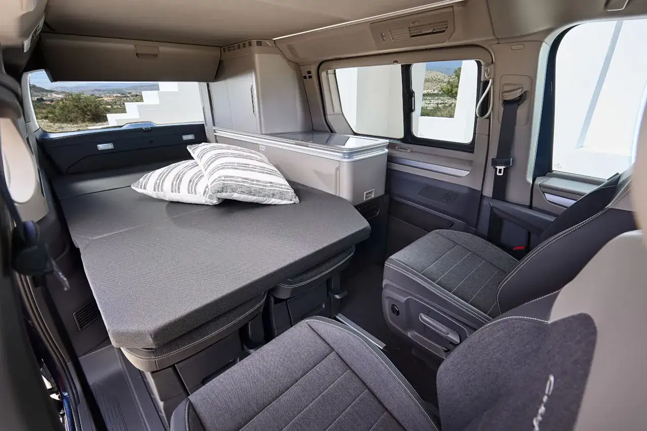 Interior de una autocaravana Volkswagen California con cama desplegada, cojines y nevera.