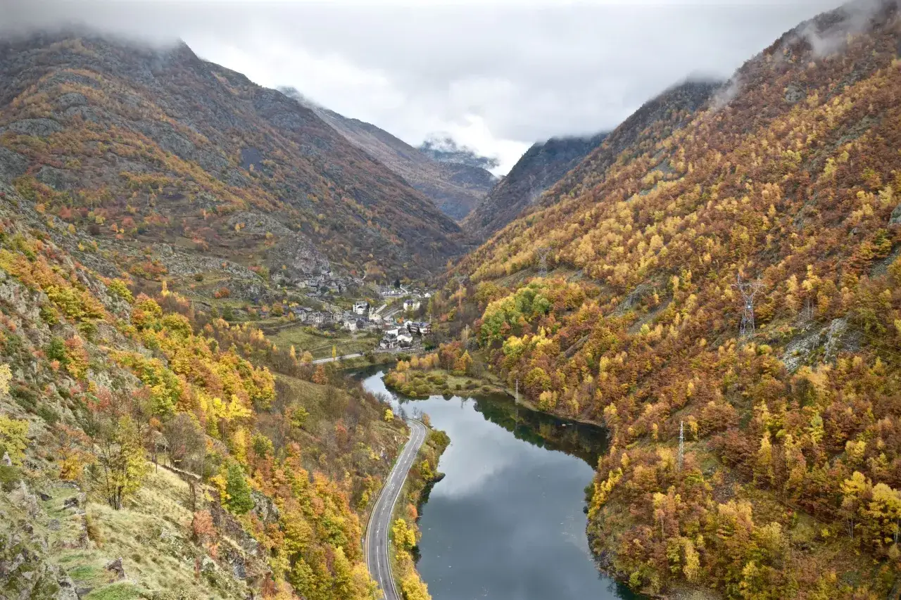 Valle otoñal con el pueblo de Esterri de Cardos, un río serpenteante y montañas cubiertas de árboles en tonos ocres y amarillos.
