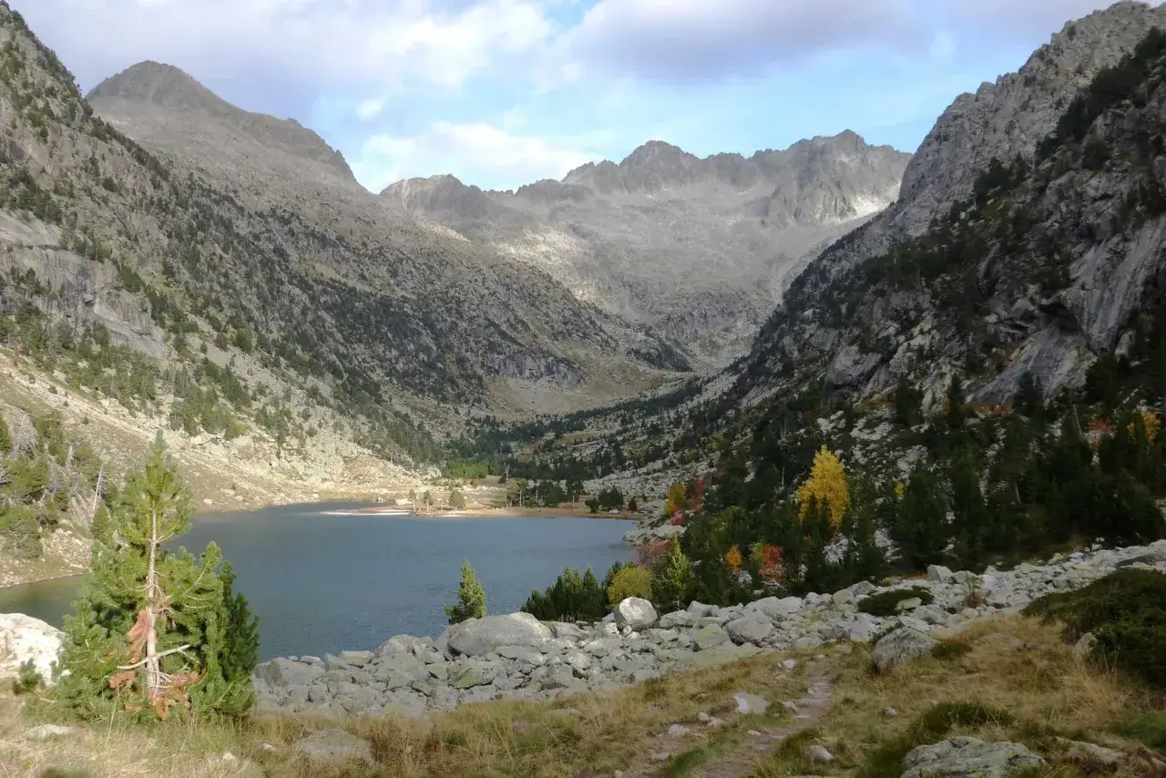 Paisaje montañoso con un lago sereno, árboles de otoño y rocas. El sol ilumina las cumbres, creando una atmósfera mágica en Besiberri Sud.
