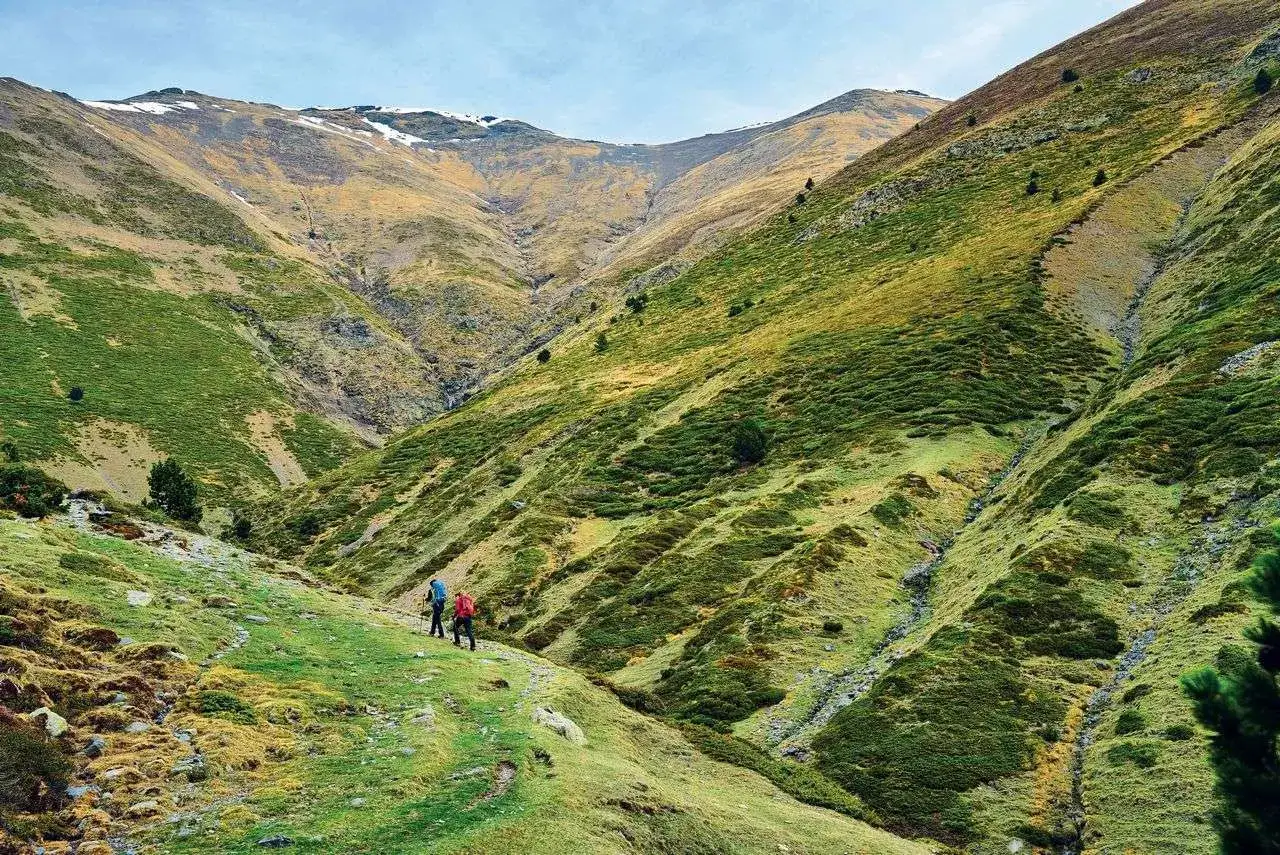 Dos excursionistas caminan por un valle verde y ondulado en el Pirineo Catalán, con montañas escarpadas y un cielo nublado.