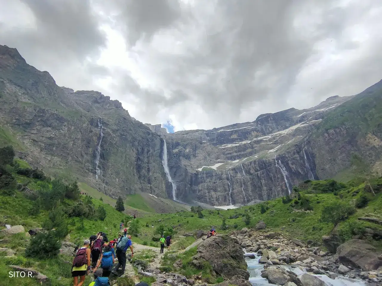 Senderismo hacia el circo de Gavarnie, con impresionantes cascadas cayendo por acantilados rocosos bajo un cielo nublado.