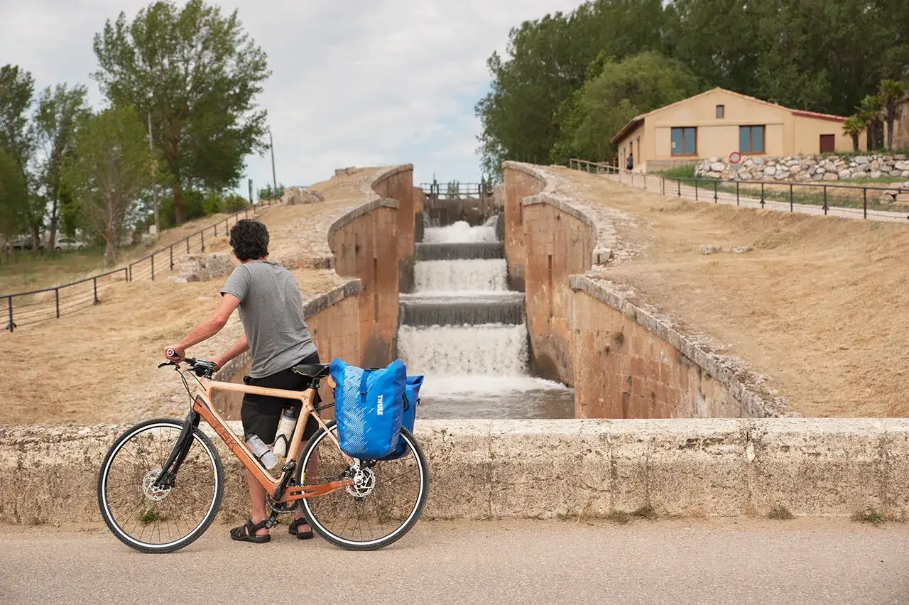 Ciclista con alforjas azules se detiene junto a una cascada en la ruta del Canal de Castilla.