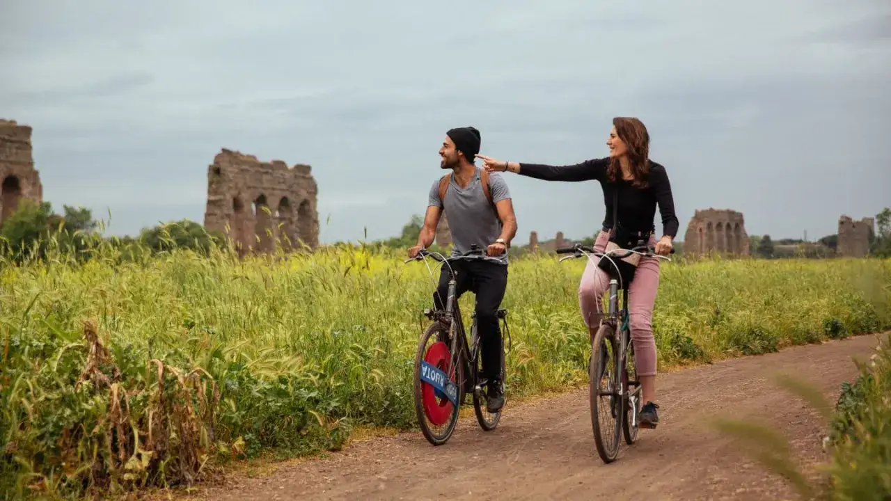 Pareja disfrutando de un paseo en bicicleta por Roma, con ruinas antiguas de fondo.