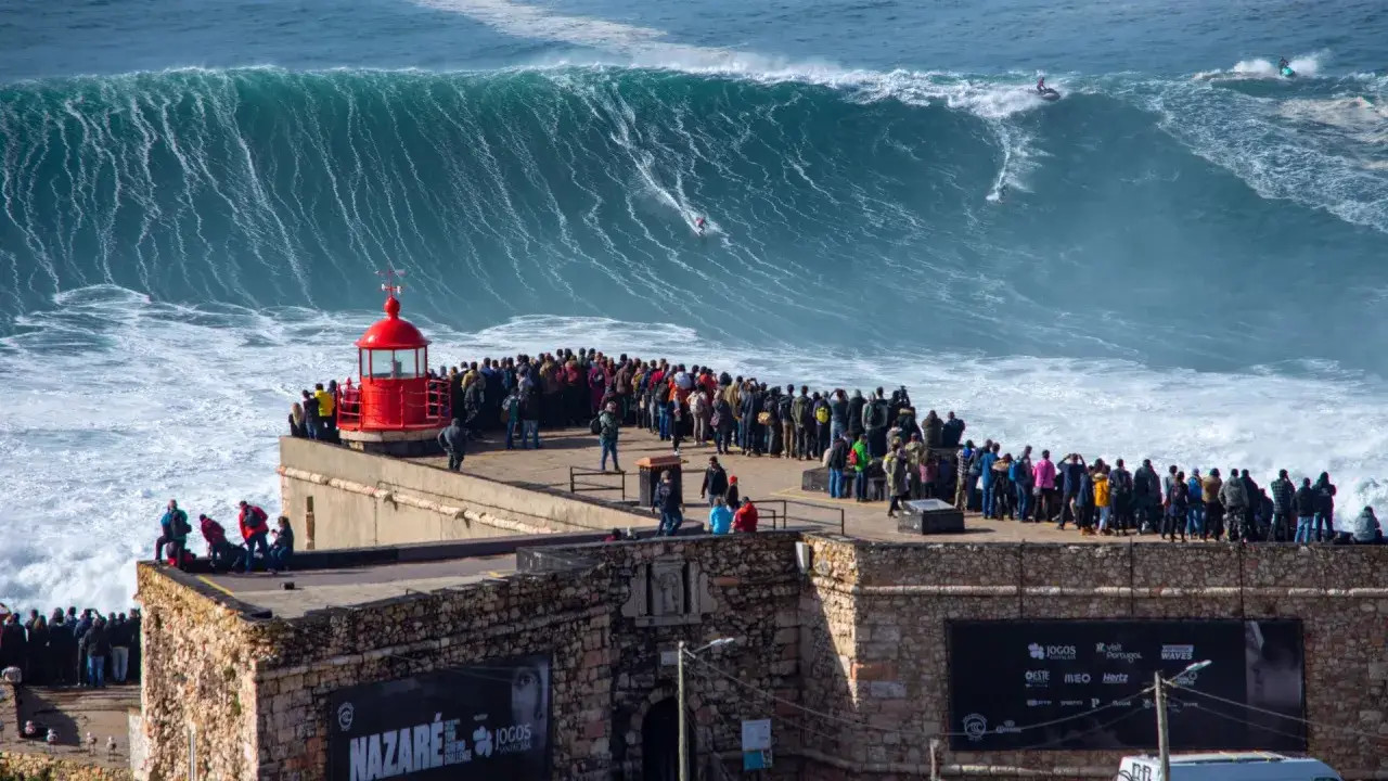 Espectadores observan surfistas desafiar las gigantes olas de Nazaré desde un acantilado. Un faro rojo destaca entre la multitud.