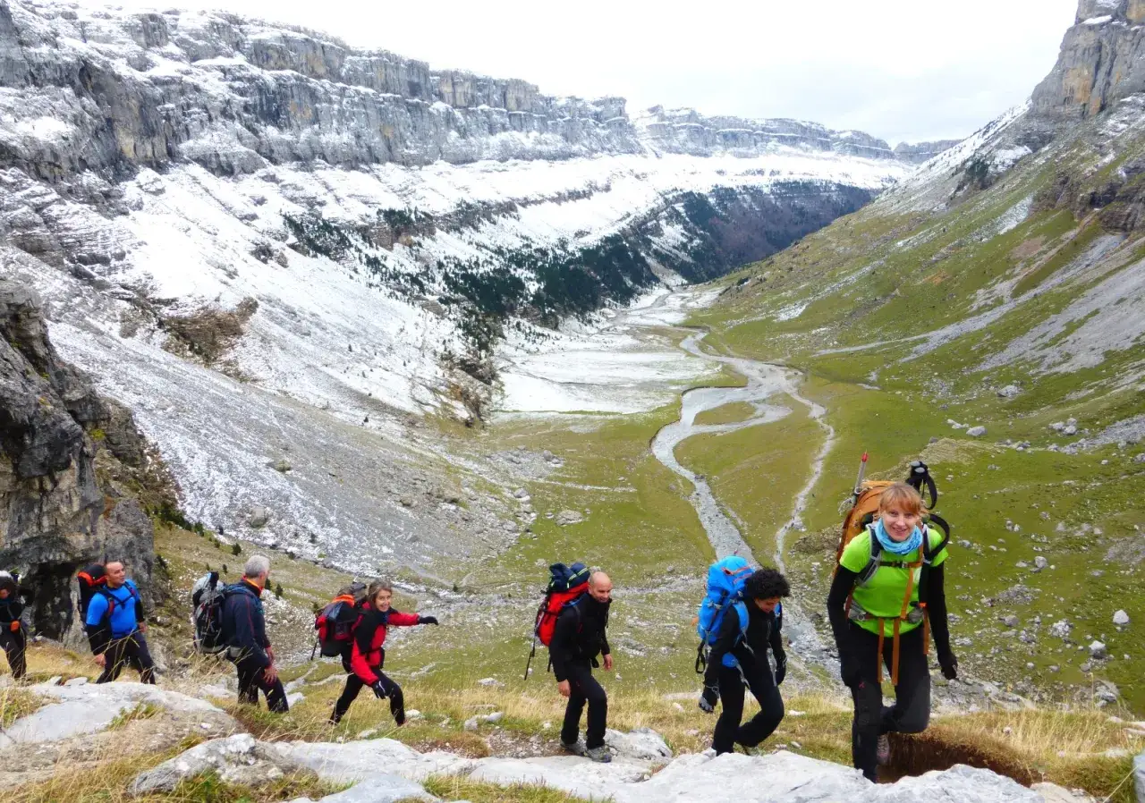 Senderismo en la pradera de Ordesa, con un río serpenteante y montañas nevadas al fondo. Un grupo de excursionistas avanza por el sendero.