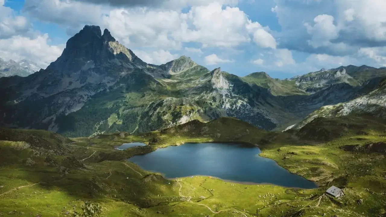 Majestuoso pico y lago azul en una ruta por los Pirineos franceses. Senderos serpentean por verdes praderas, con tiendas de campaña y una cabaña.