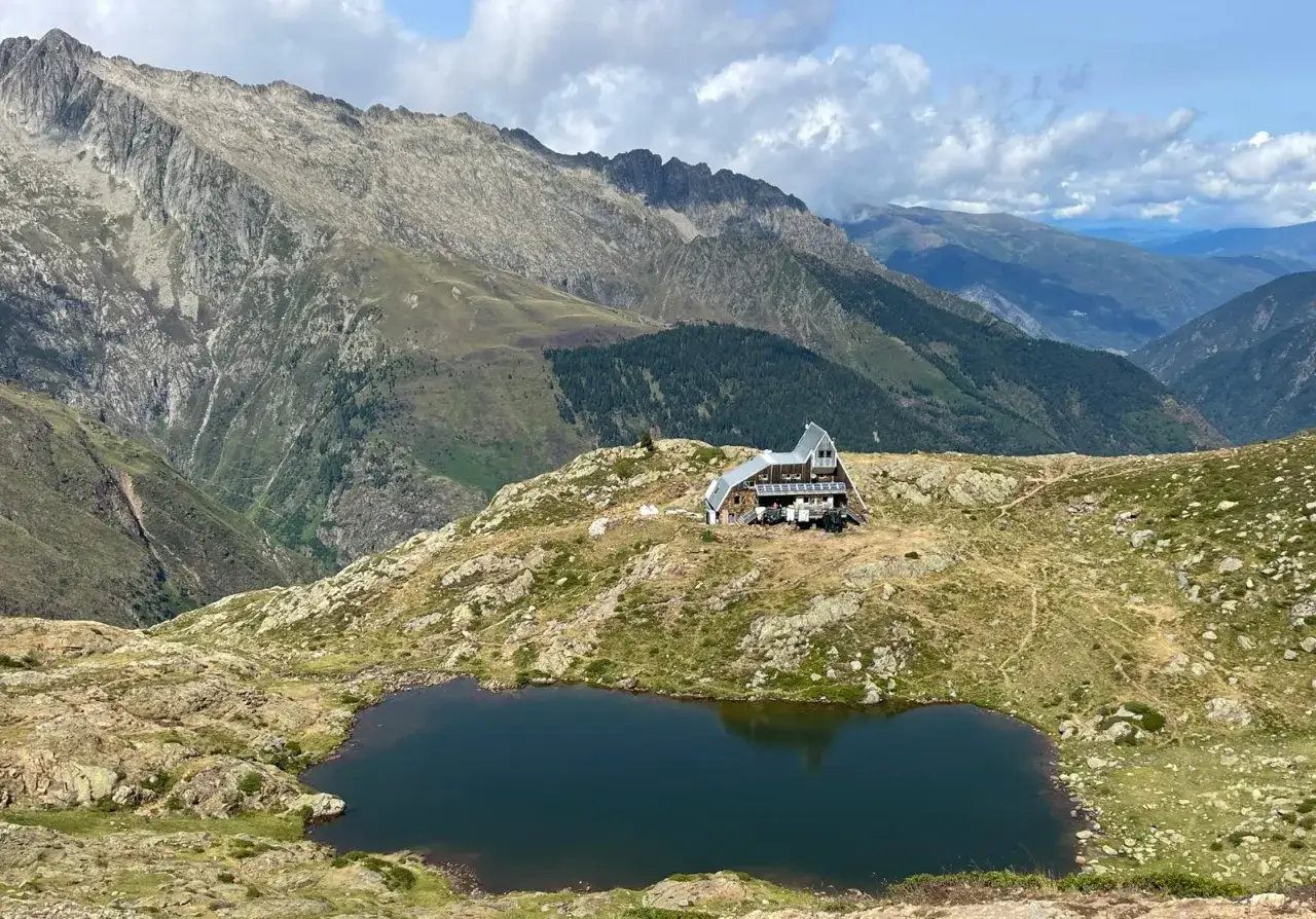 El refugio de Vallferrera se alza en una ladera rocosa junto a un lago de montaña, con imponentes picos nevados al fondo.