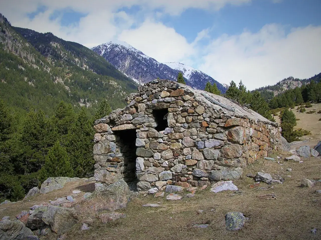 Antigua cabaña de piedra en el Vall de la Llosa, rodeada de pinos y montañas nevadas.