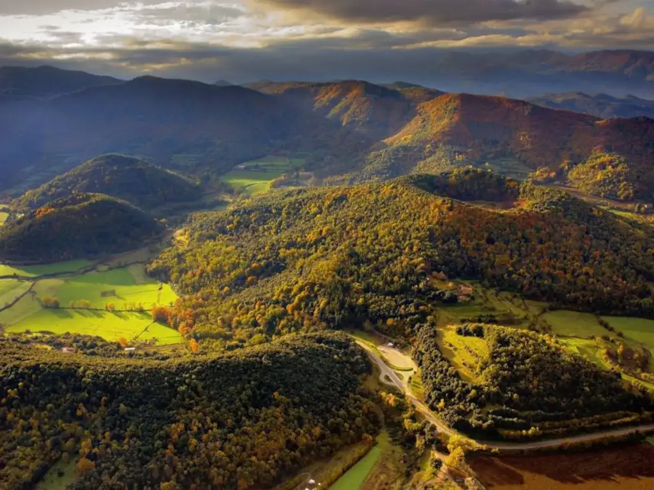 Paisaje otoñal en La Garrotxa, con colinas cubiertas de bosques de colores cálidos y campos verdes bajo un cielo nublado.