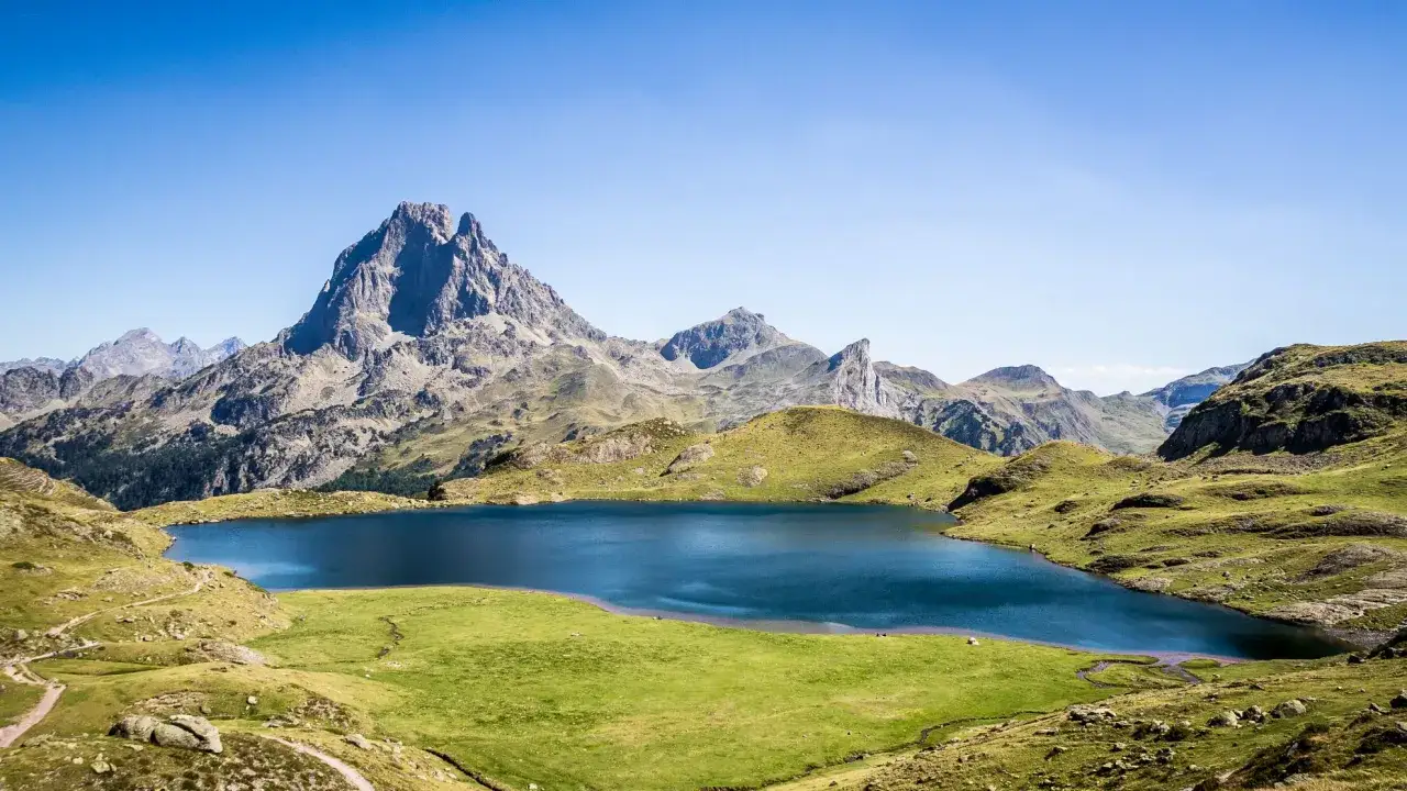 Lago azul sereno en el Valle de Ossau, rodeado de verdes colinas y picos montañosos imponentes bajo un cielo despejado.