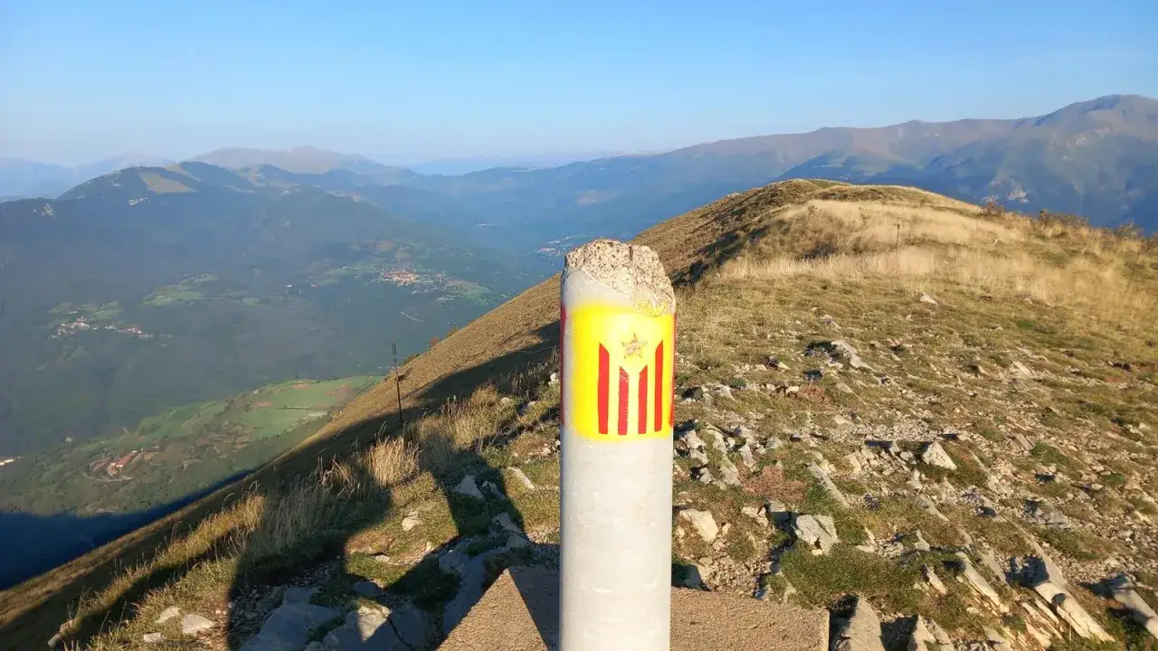 Un poste de cemento con la bandera catalana, el taga, marca el camino en una cresta montañosa con vistas a un valle verde.