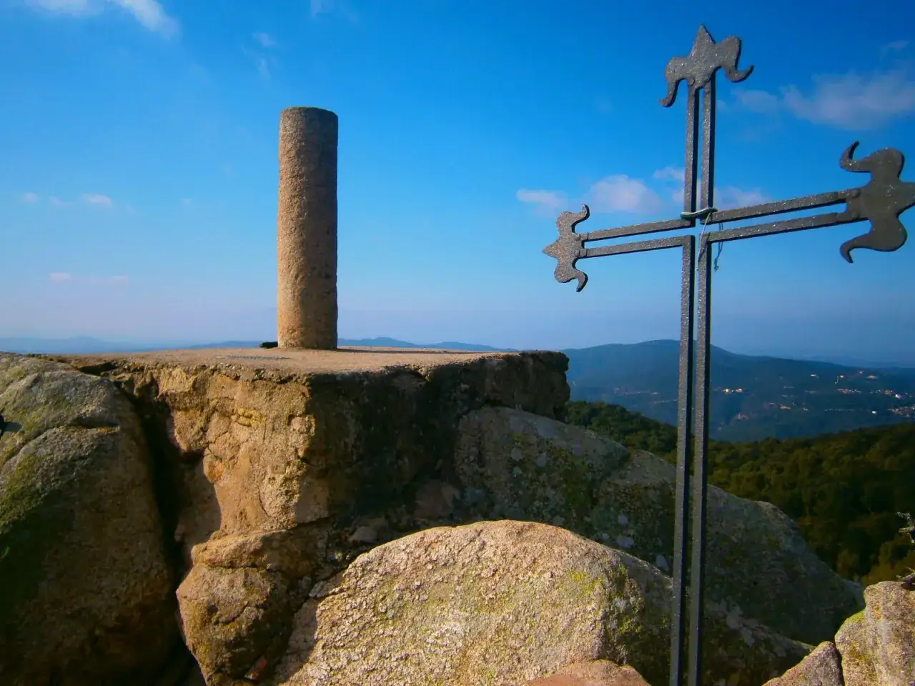 Cruz de hierro y pilar de piedra en el Puig de Cadiretes, con vistas a un paisaje montañoso y el cielo azul.