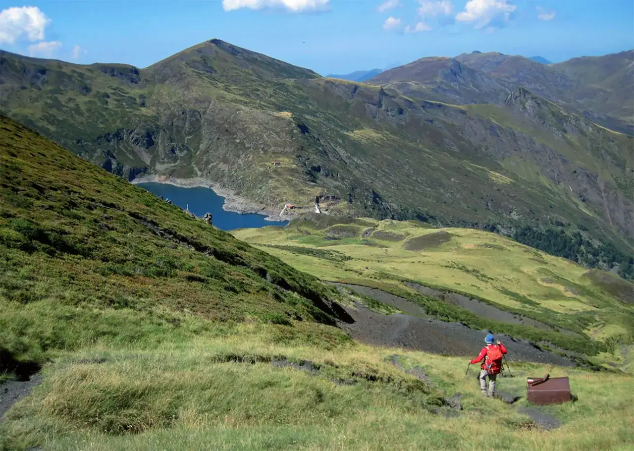 Senderista con mochila roja desciende por un sendero en las montañas de la Val d'Aran, con un lago azul al fondo.
