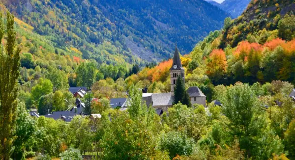 Pueblo en el **Valle de Arán** rodeado de árboles con hojas de otoño en tonos verdes, amarillos y naranjas. Destaca una iglesia con campanario.