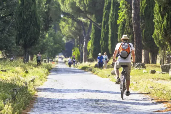 Un hombre pedalea por un camino empedrado en Roma, rodeado de altos cipreses y ruinas antiguas.